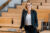 Zu sehen ist eine Frau mit einer Brille in einem Hörsaal, die in die Kamera lächelt und sich an einen Schreibtisch lehnt.