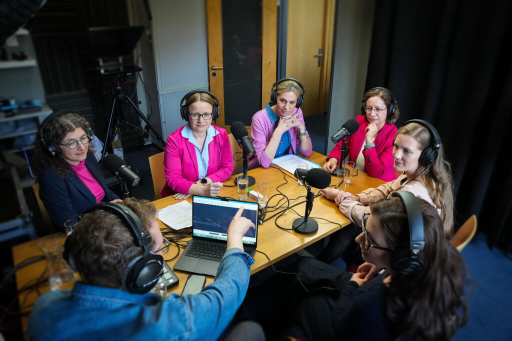 Zu sehen sind die Sprecher:innen des Podcasts recht:zeitig, versammelt an einem Tisch vor Mikrofonen